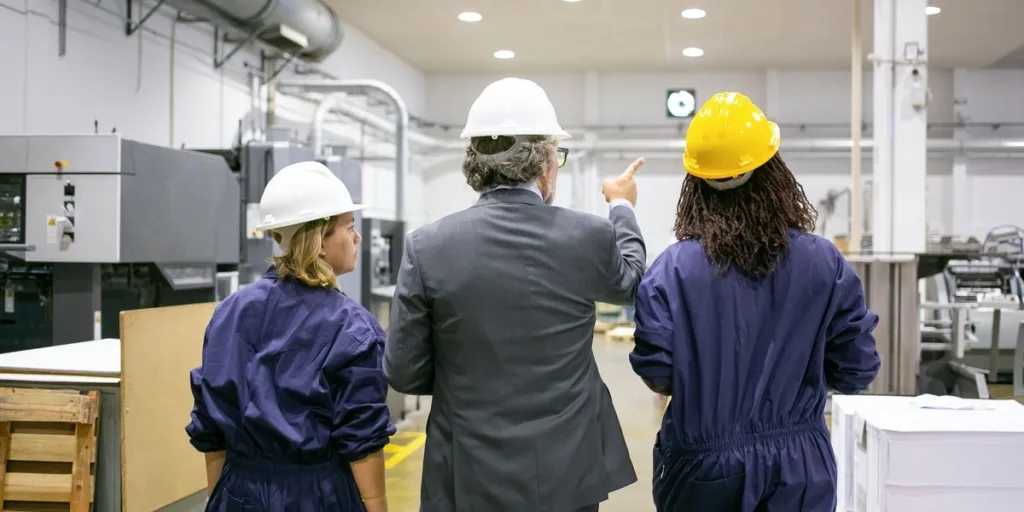 ingeniero masculino empleadas fabrica cascos caminando sobre piso planta hablando hombre apuntando al equipo e instruyendo mujeres